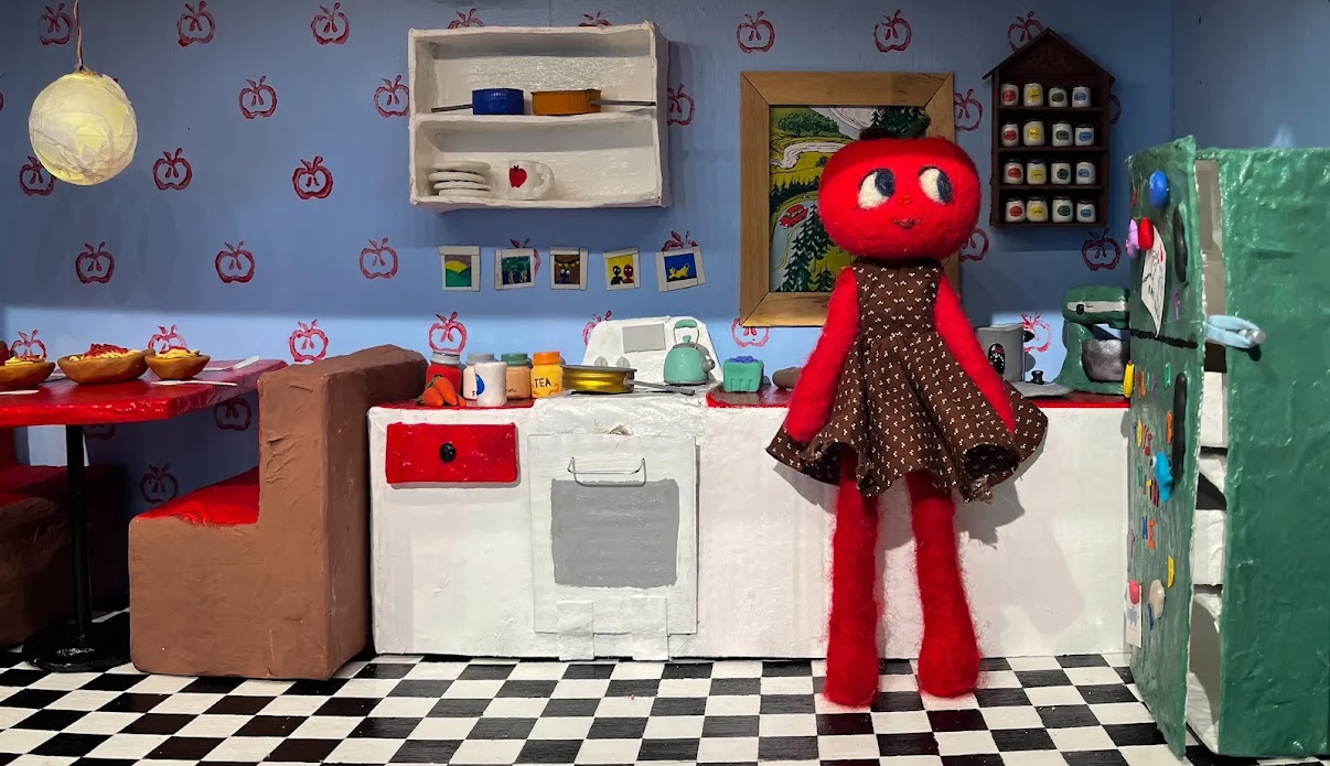 A handmade doll stands in the middle of a dollhouse kitchen. The doll is made out of red felt and has a head that looks like a apple.